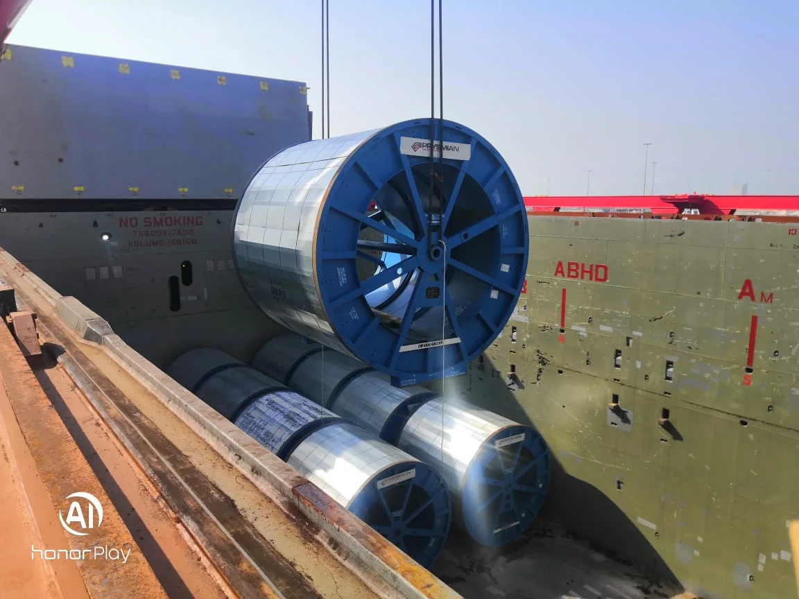 Large cable drum suspended over steel coils in a ship hold.