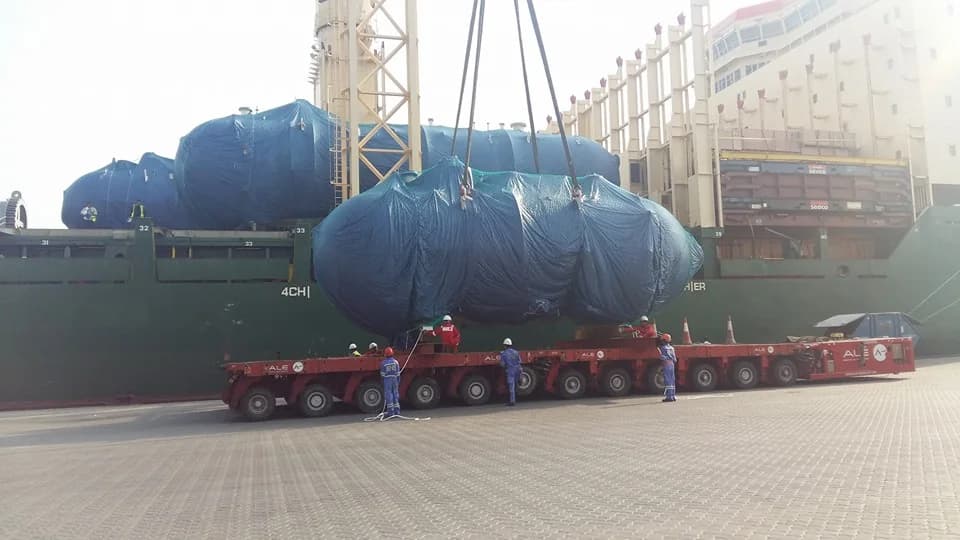 Covered industrial cargo on a transport trailer beside a ship.