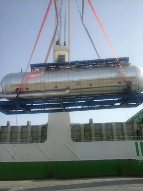 Long cylindrical industrial cargo being lifted beside a vessel.