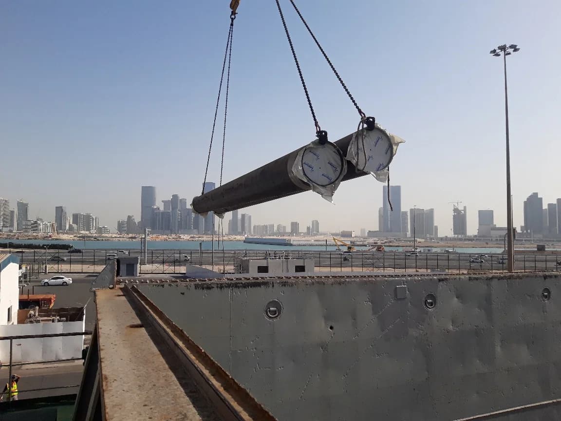 Two long pipes suspended by crane above a vessel hold.