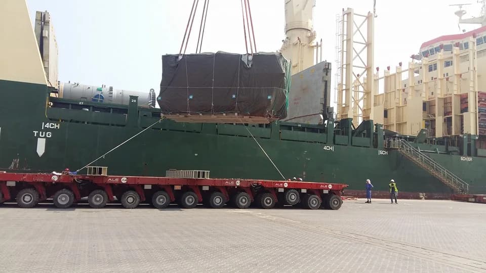 Covered rectangular cargo suspended over a multi-axle trailer beside a vessel.