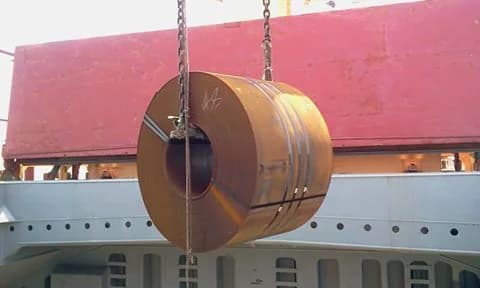 Large steel coil suspended by crane above a transport trailer.